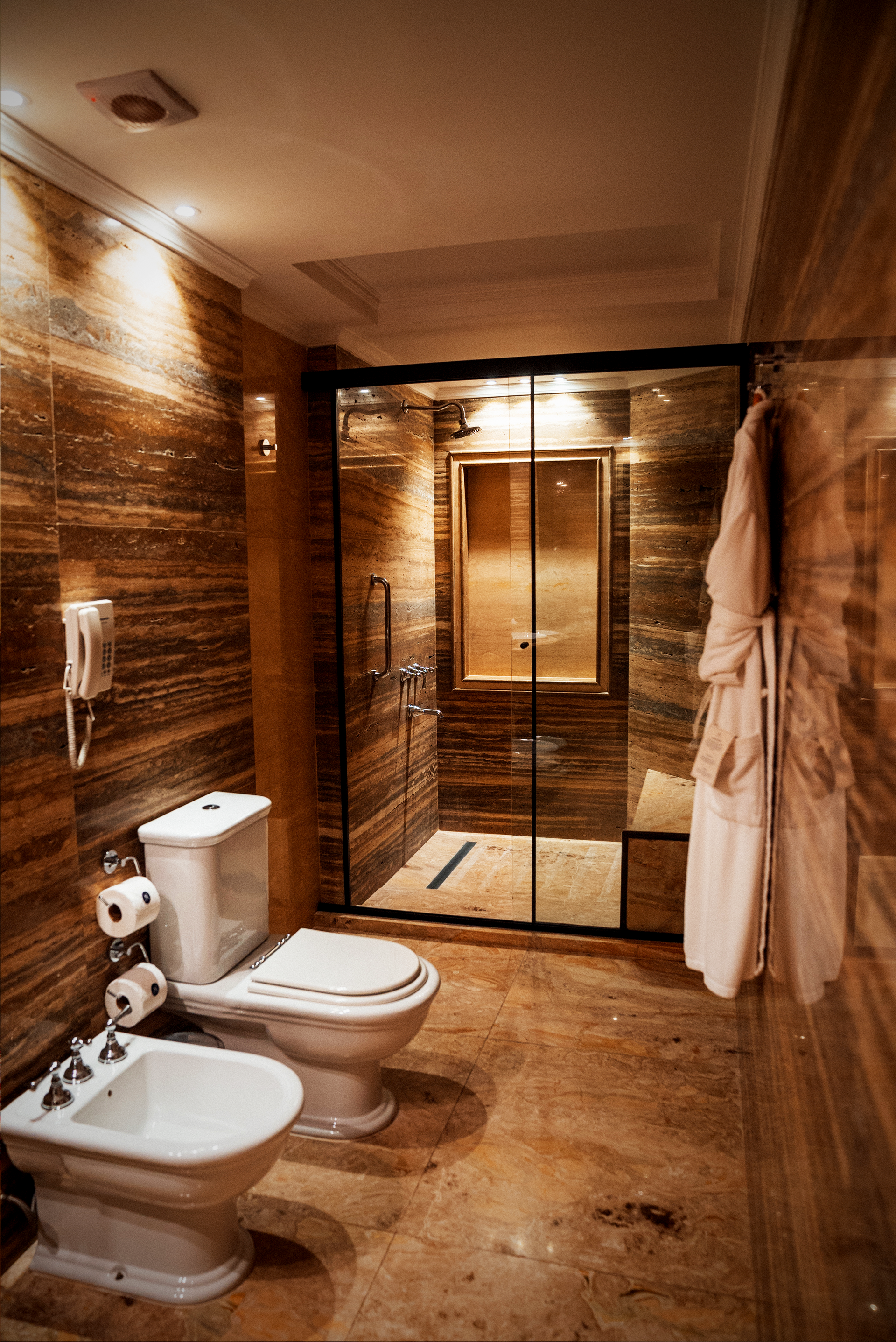 Un baño moderno con paredes de piedra marrón, lavabo blanco, bidé, inodoro y ducha con mampara de cristal y albornoz.