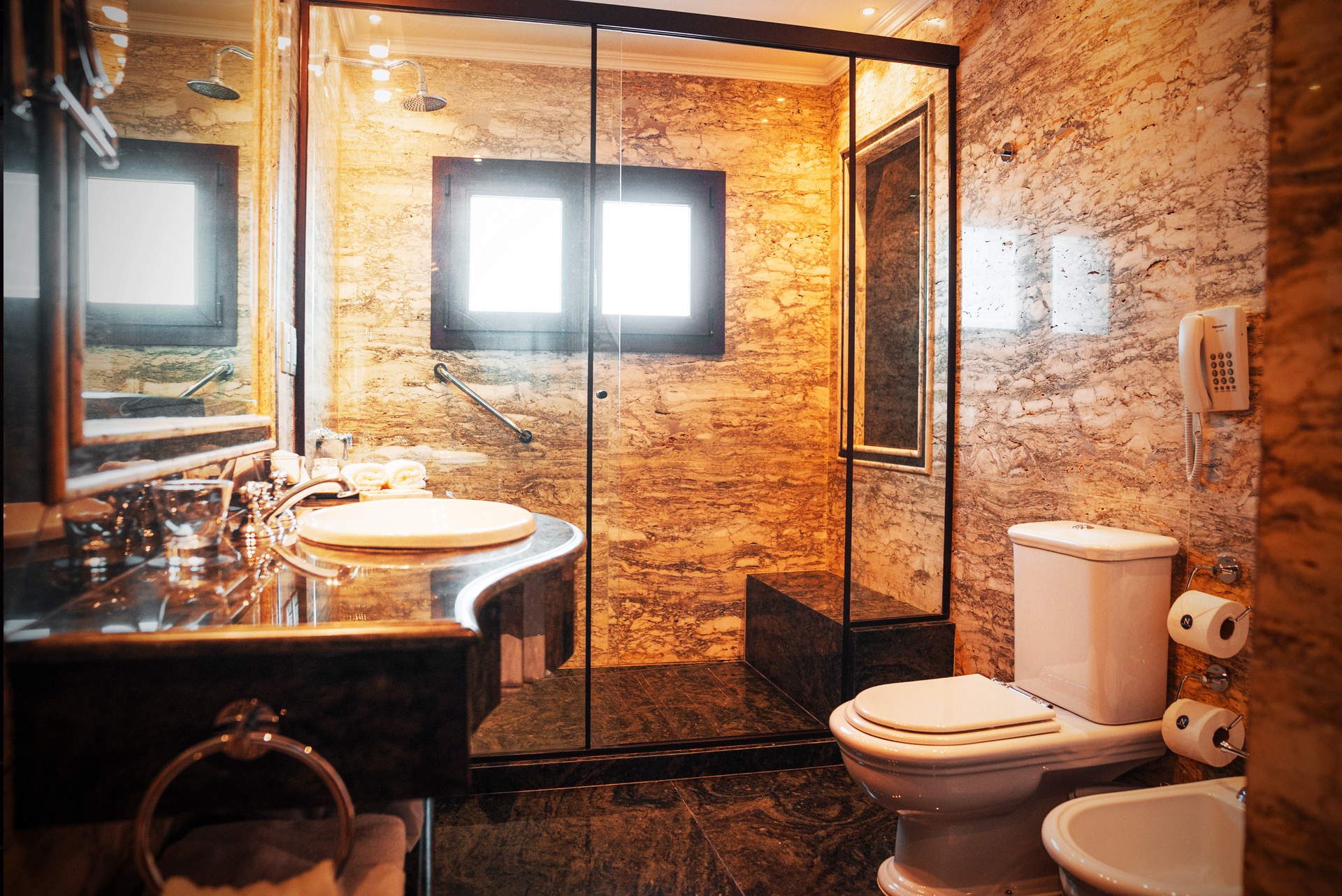 Baño moderno con paredes de mármol color canela, ducha con mampara de cristal, inodoro blanco, bidé y tocador de mármol oscuro.