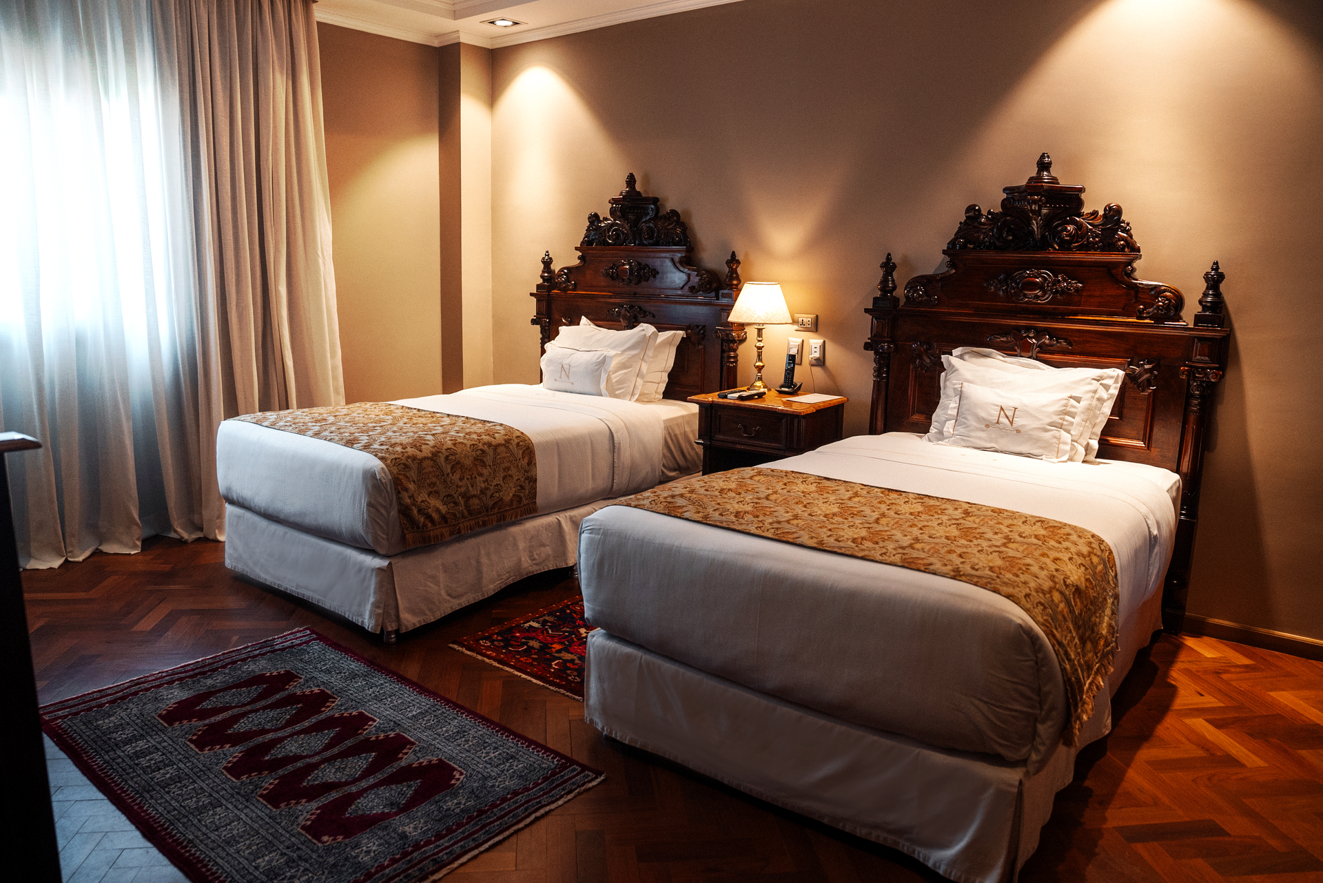 Una habitación de hotel con dos camas, cabeceros antiguos de madera tallada, ropa de cama de color tostado, una mesita de noche central y cortinas beige.