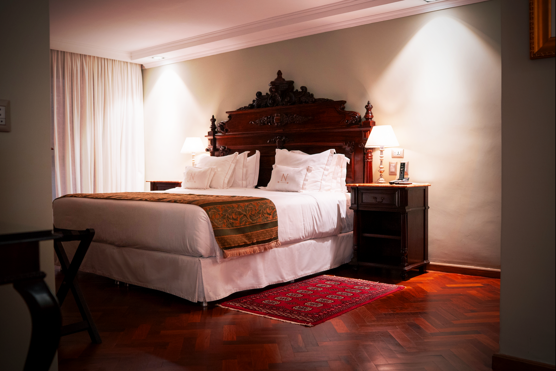 Primer plano de una cama bien hecha con almohadas y ropa de cama blancas, situada junto a una mesita de noche de madera con una lámpara encendida.