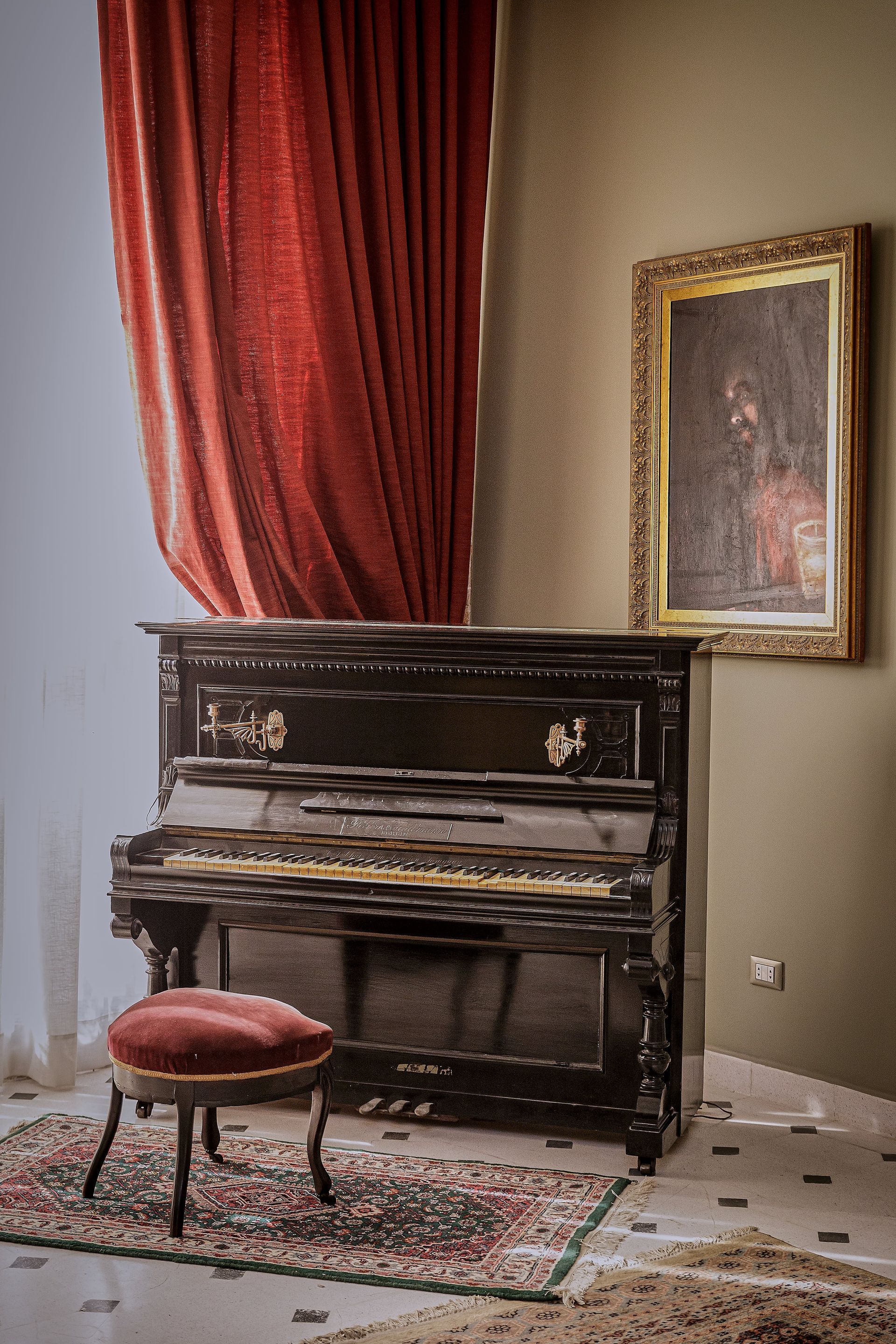 Piano vertical con taburete, cortina roja, obra de arte enmarcada y alfombra estampada en una habitación.