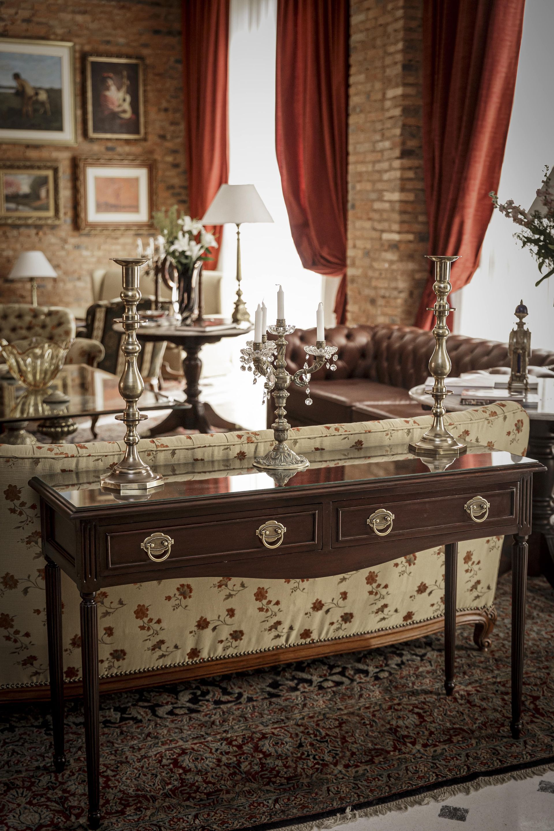 Sala de estar con muebles antiguos, pared de ladrillo, cortinas rojas y obras de arte.