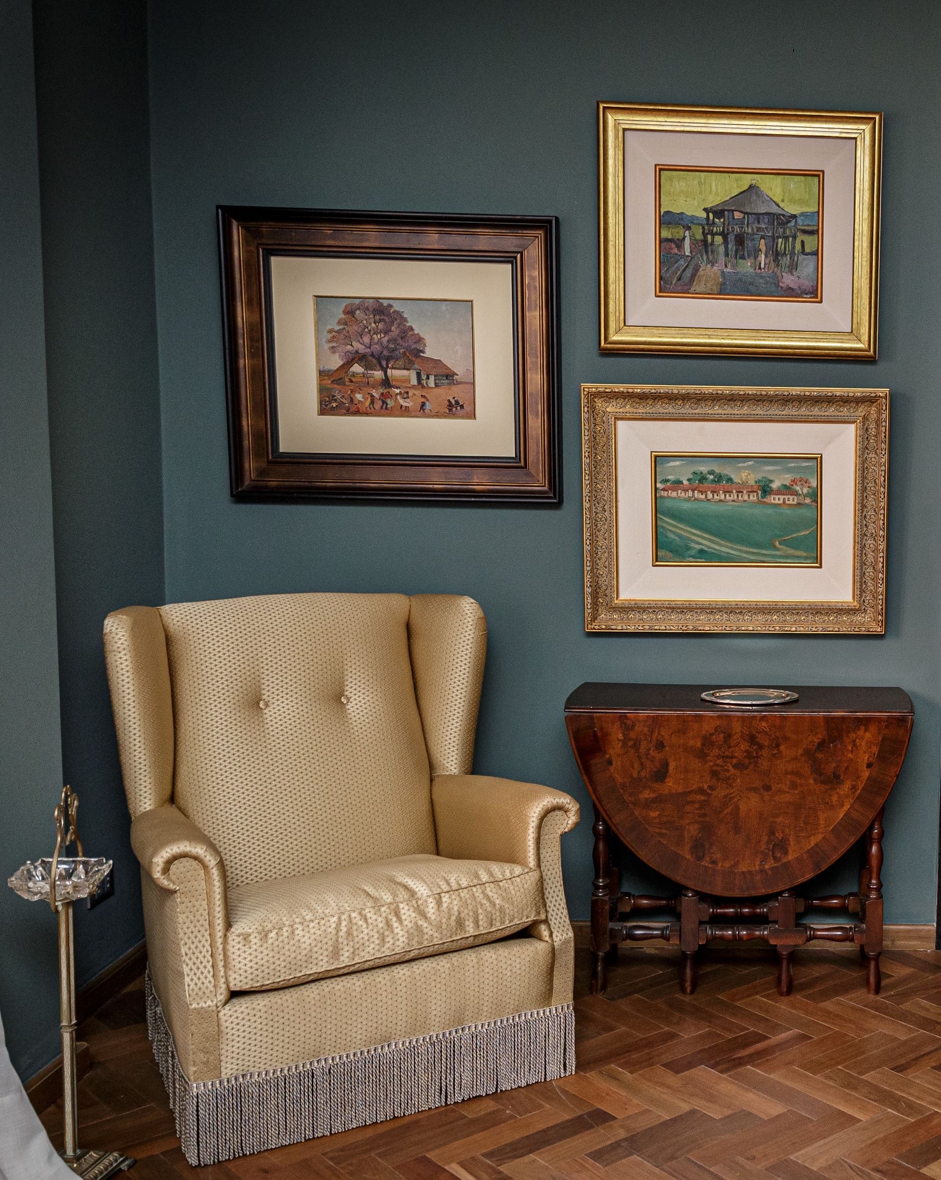 Habitación de esquina con un sillón con flecos, una mesa antigua, tres cuadros enmarcados y un soporte de plata. 