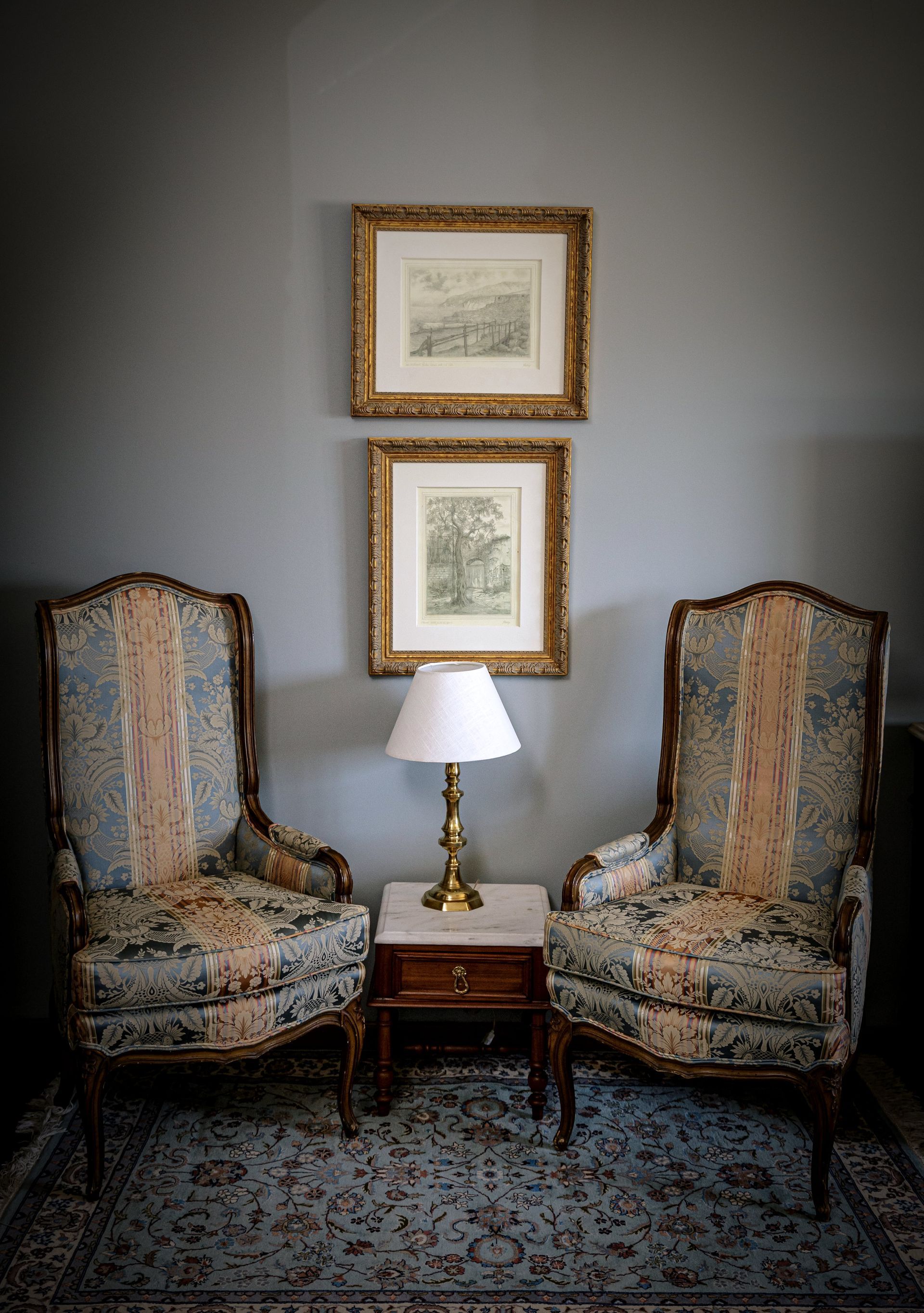 Dos sillones ornamentados flanquean una mesita con lámpara y dos láminas de paisajes enmarcadas. 