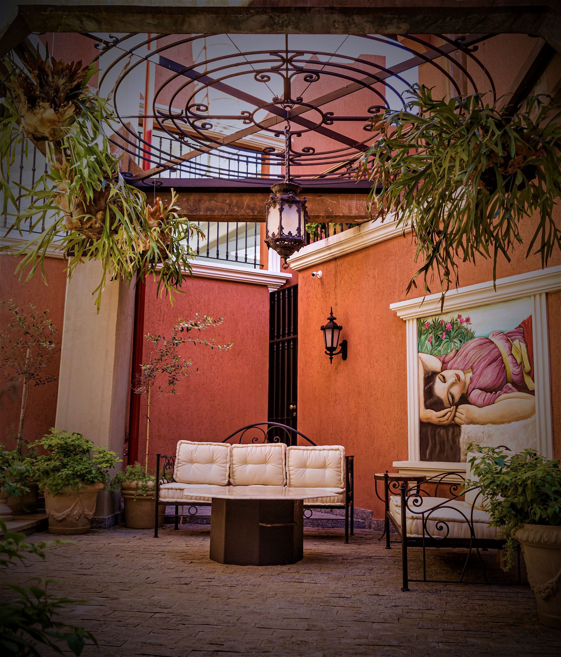 Patio con asientos, plantas colgantes, herrajes y un cuadro en la pared. Muros rojizos rodean el espacio.