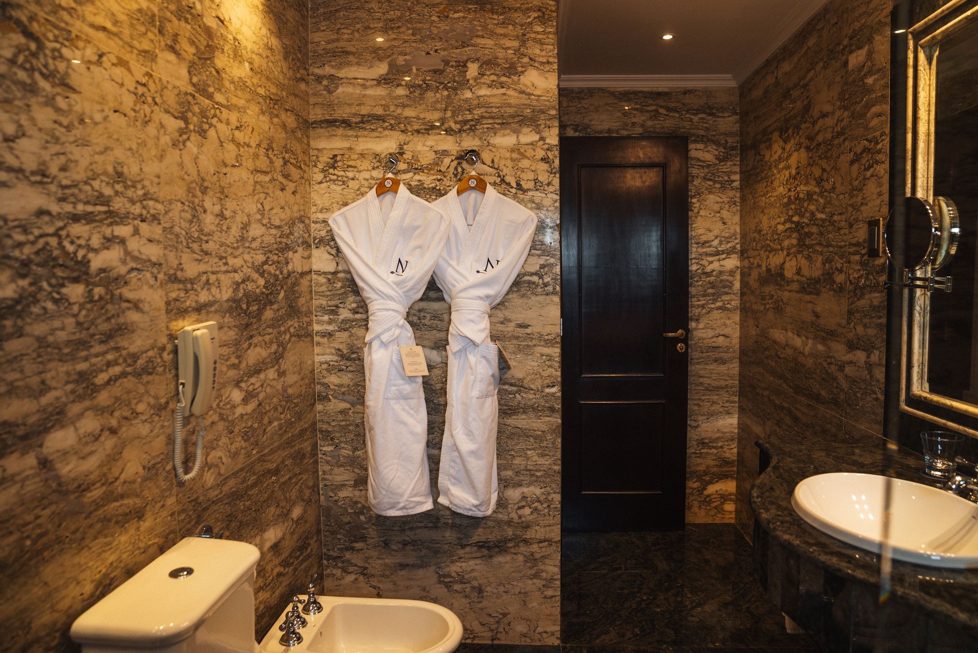 Un baño de hotel con paredes de mármol que cuenta con dos albornoces blancos, un bidé, un inodoro y un lavabo.