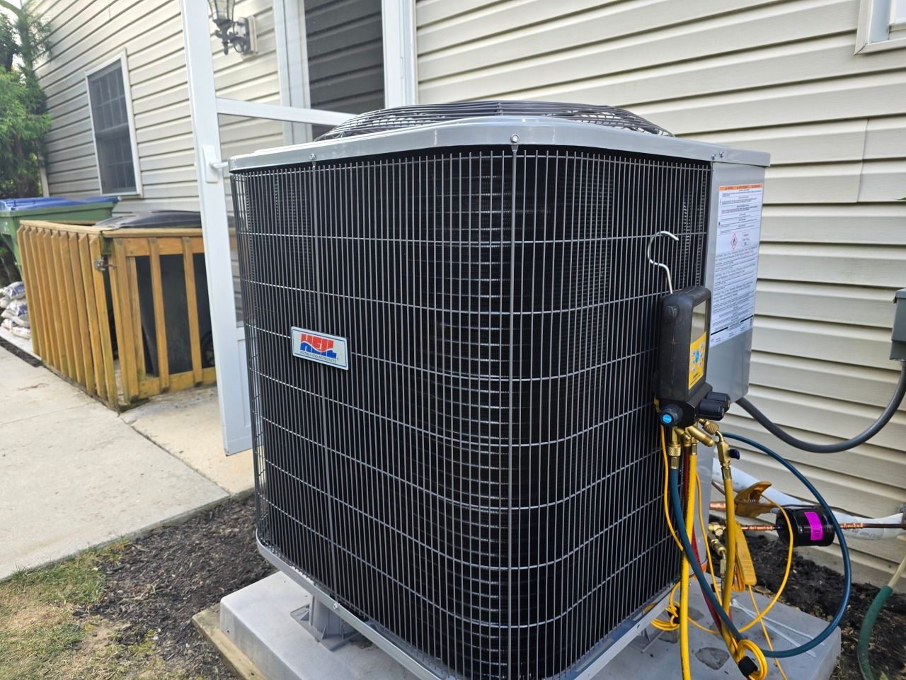 An outdoor HVAC unit sits on a concrete pad next to a house with beige siding, featuring connected diagnostic gauges.