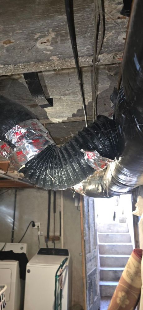 Flexible black and silver ductwork hangs from a ceiling above a basement laundry area with stairs in the background.