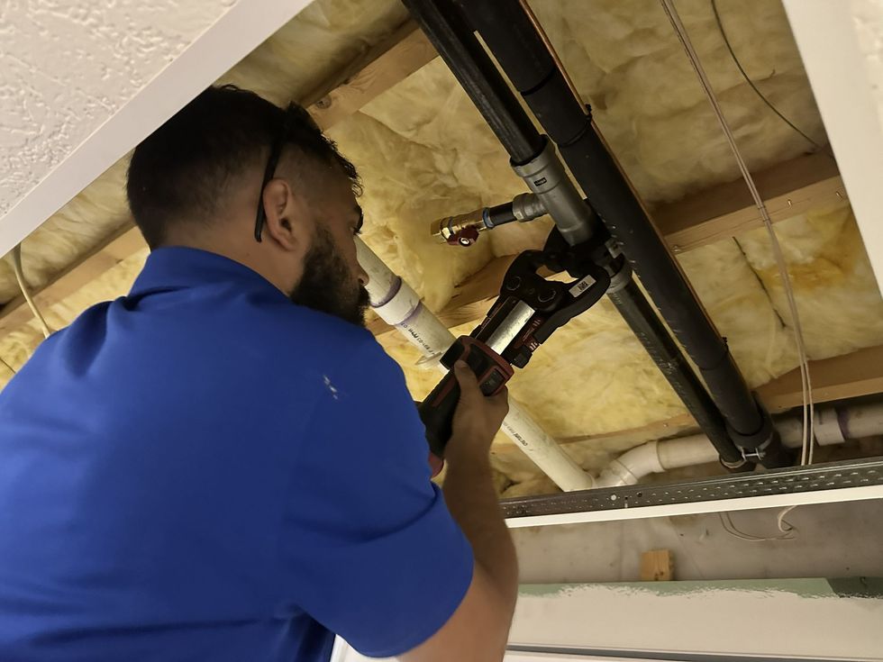 A worker in a blue shirt uses a pipe-crimping tool on metal pipes in an open ceiling with exposed insulation.
