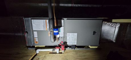 HVAC air handler unit installed in an attic with visible plumbing pipes and insulation.