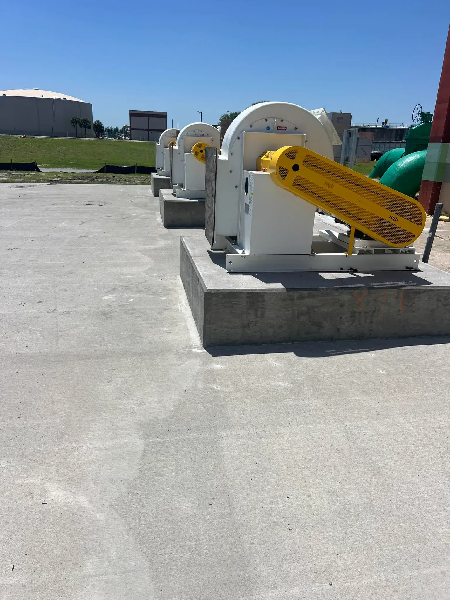 Three white centrifugal blowers with yellow safety belt guards on concrete pads in an outdoor industrial area.