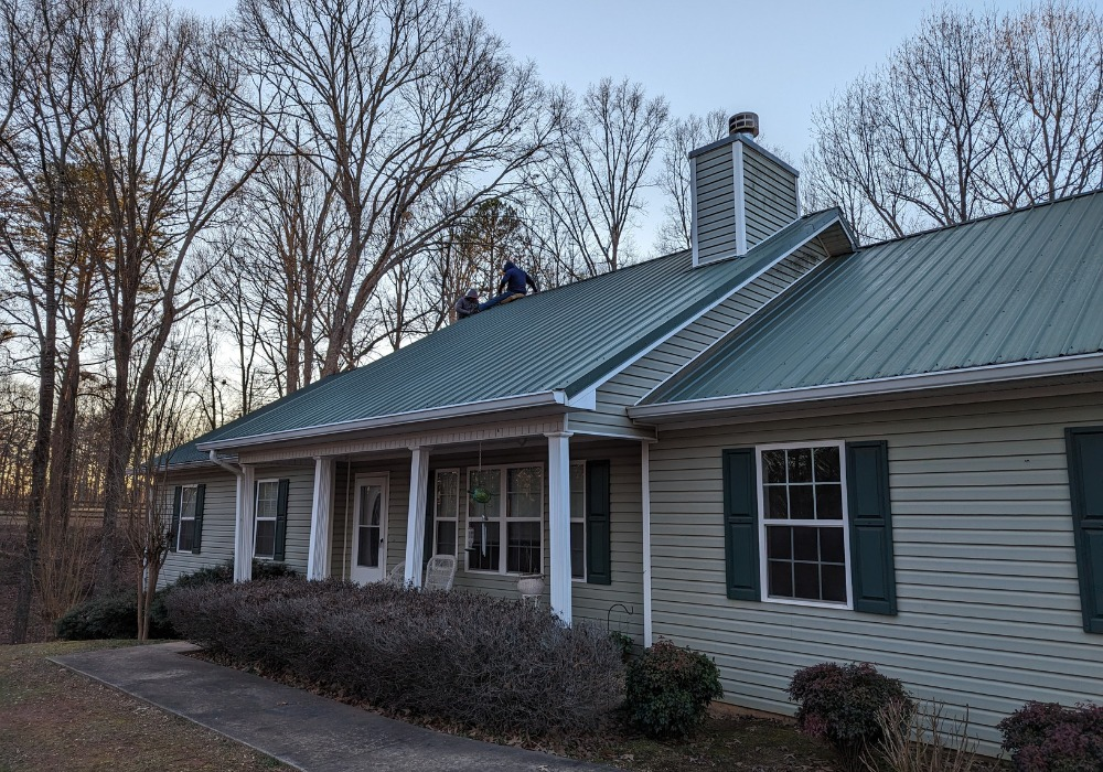 Green metal roof on a house, with a person working on top. Trees in the background.