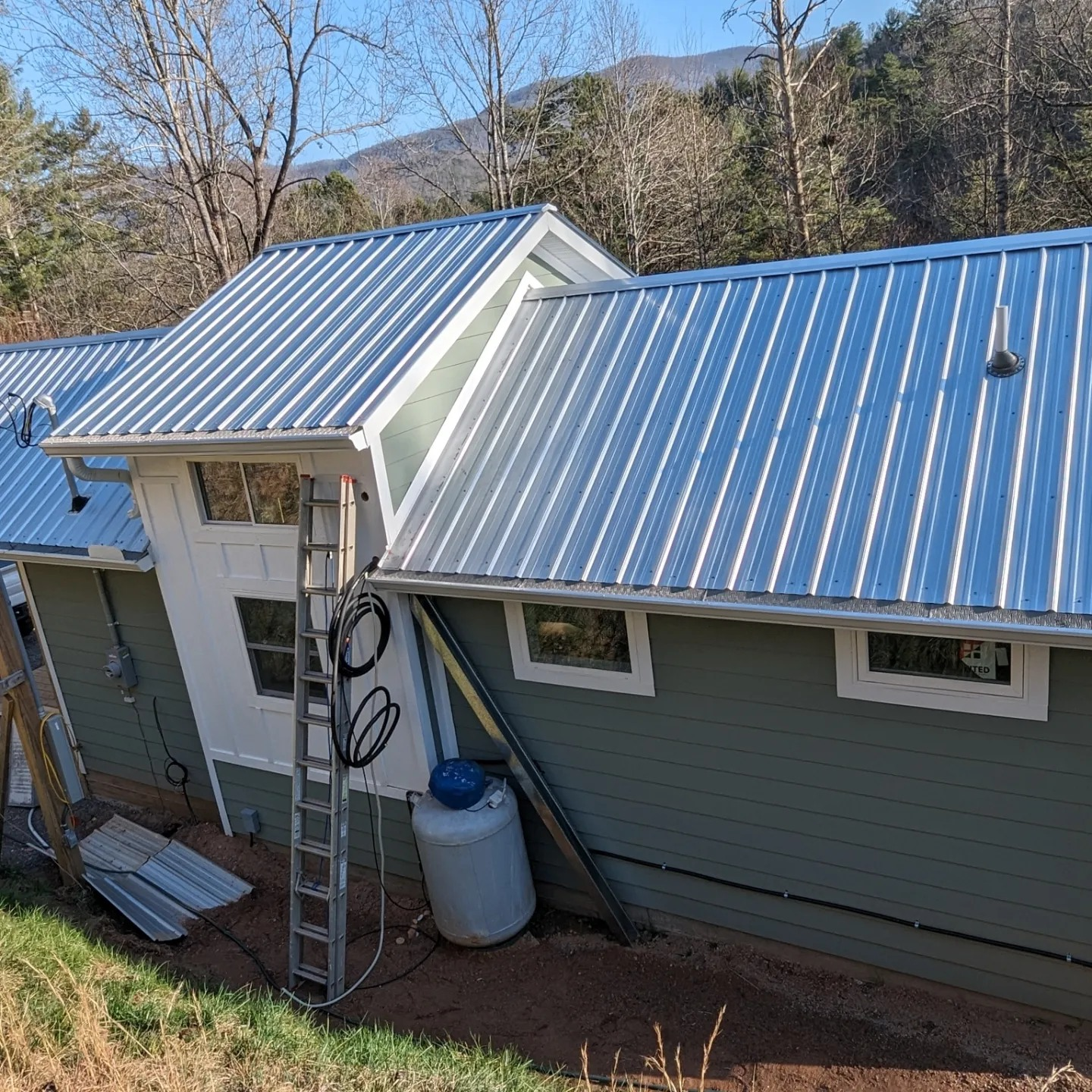 Green house with silver metal roof under construction; ladder, propane tank, and mountains visible.