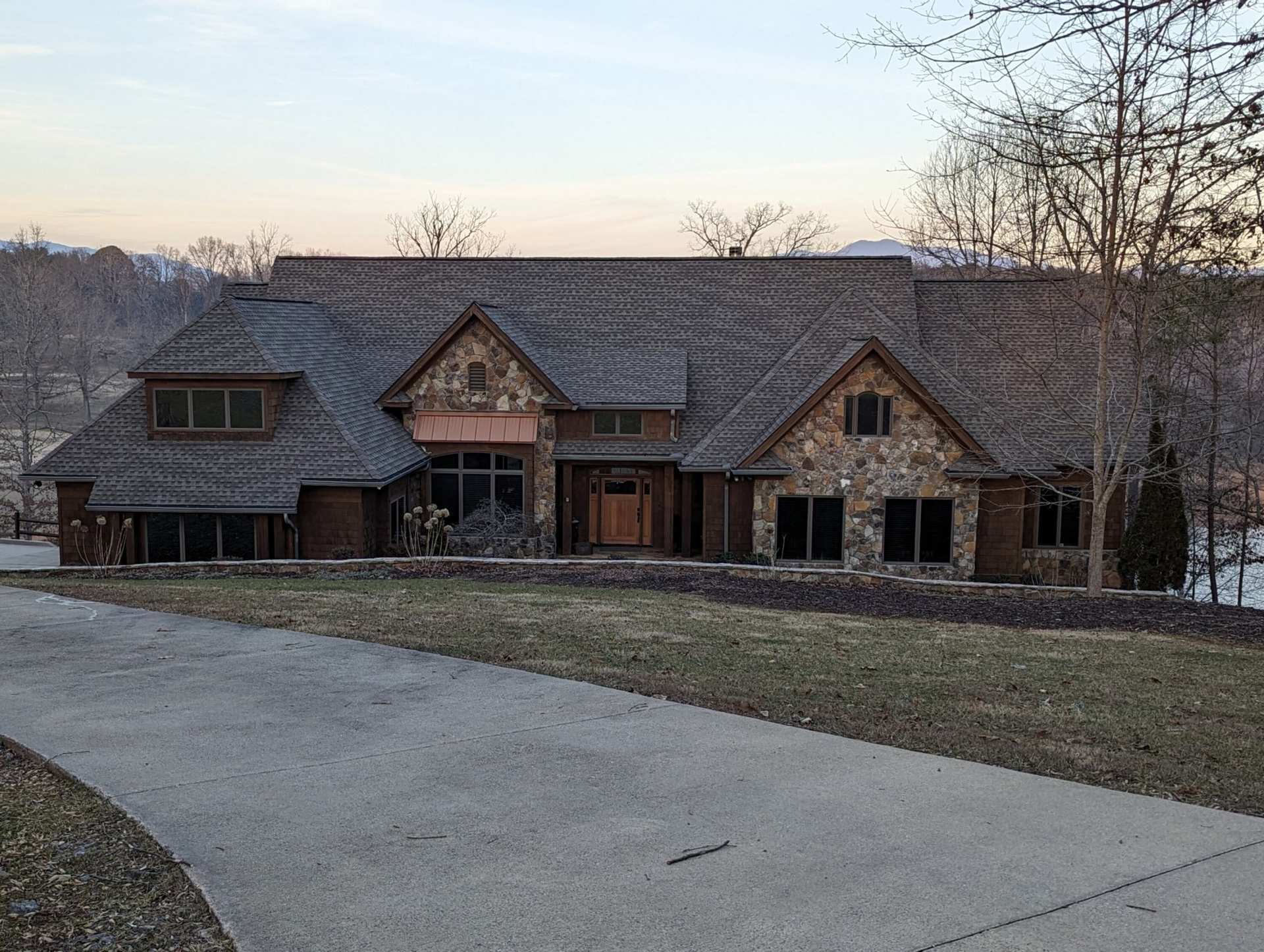 Stone and wood-sided house with a long driveway and dark roof. Trees and a slightly overcast sky are in the background.