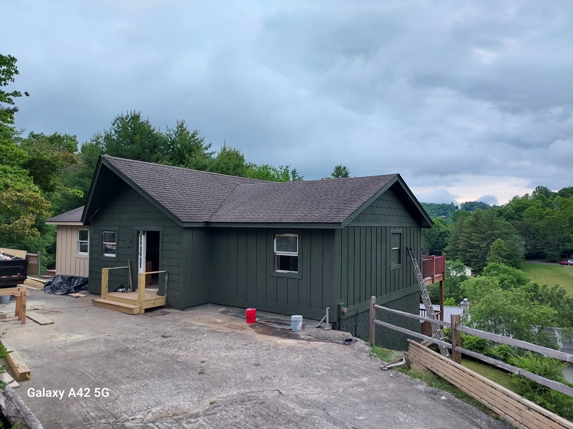 Green-sided house with brown roof under overcast sky; construction materials and partial wooden fence in view.