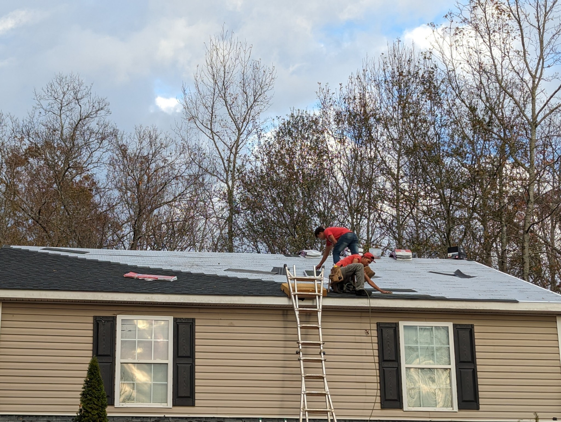Two roofers repairing a roof on a beige house; a ladder is positioned on the roof; trees and cloudy sky in the background.