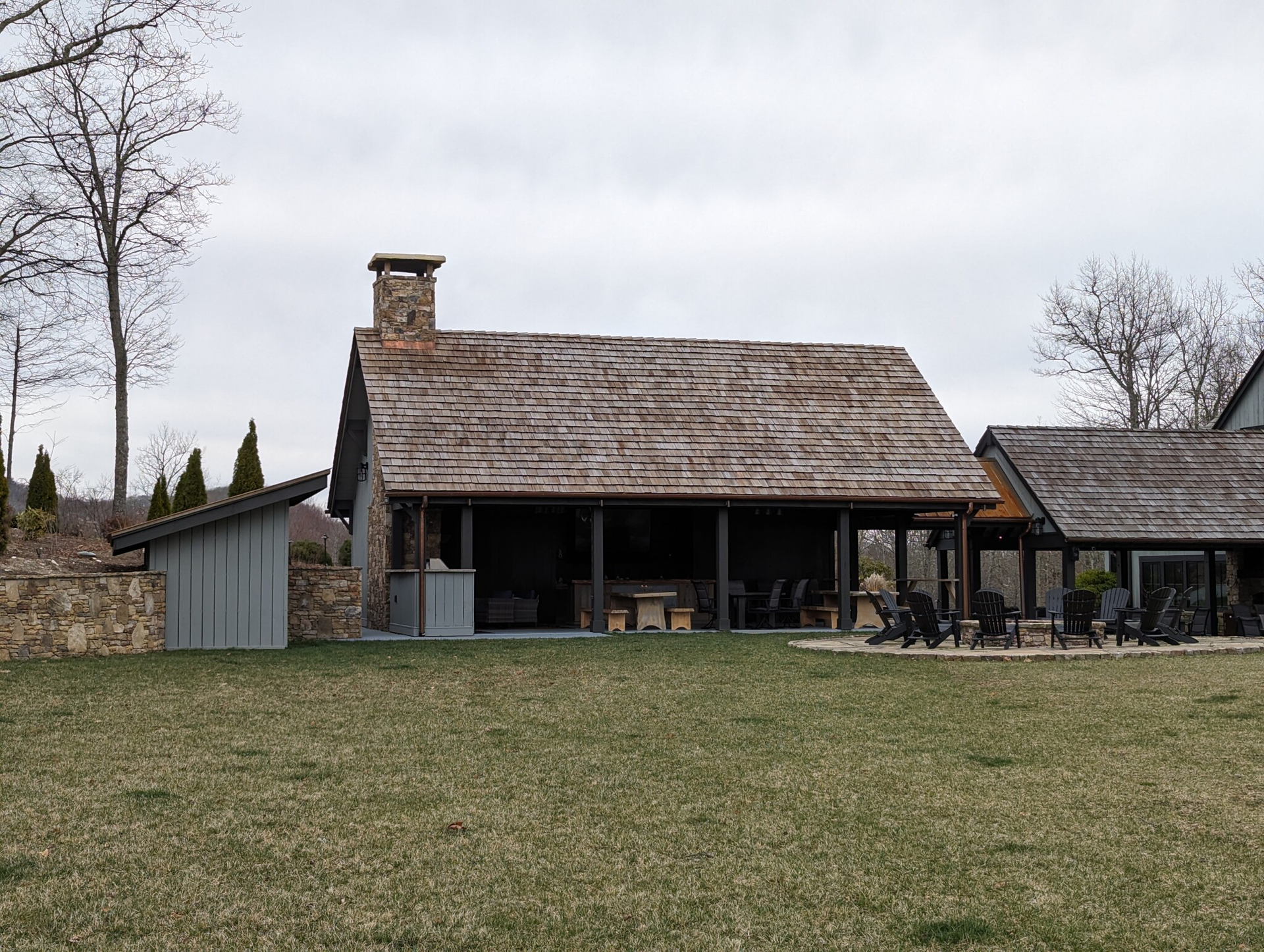 Stone cabin with wooden shingle roof, chimney, and covered porch, set on a grassy lawn under an overcast sky.
