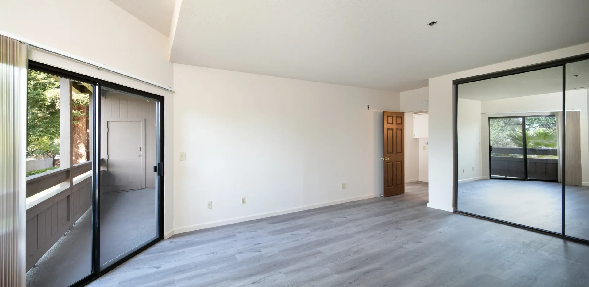 Empty apartment bedroom with wood-style flooring, mirrored closet doors, and sliding glass doors leading to a balcony.