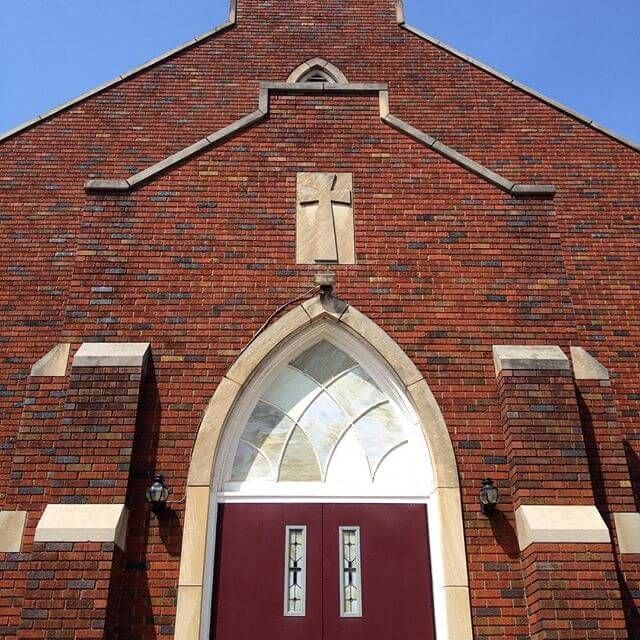 Front facade of a red brick church with a pointed arched entrance and maroon double doors.