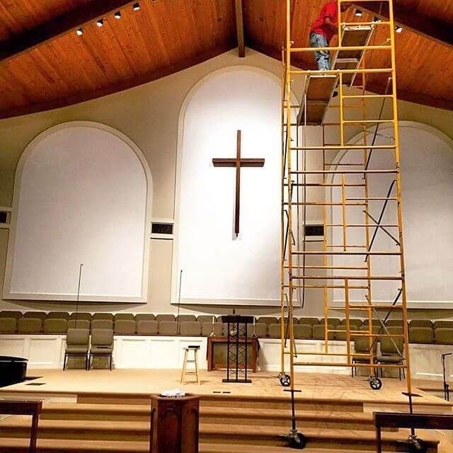 Church sanctuary with a large cross on the wall and a tall ladder beside the altar under a wooden ceiling