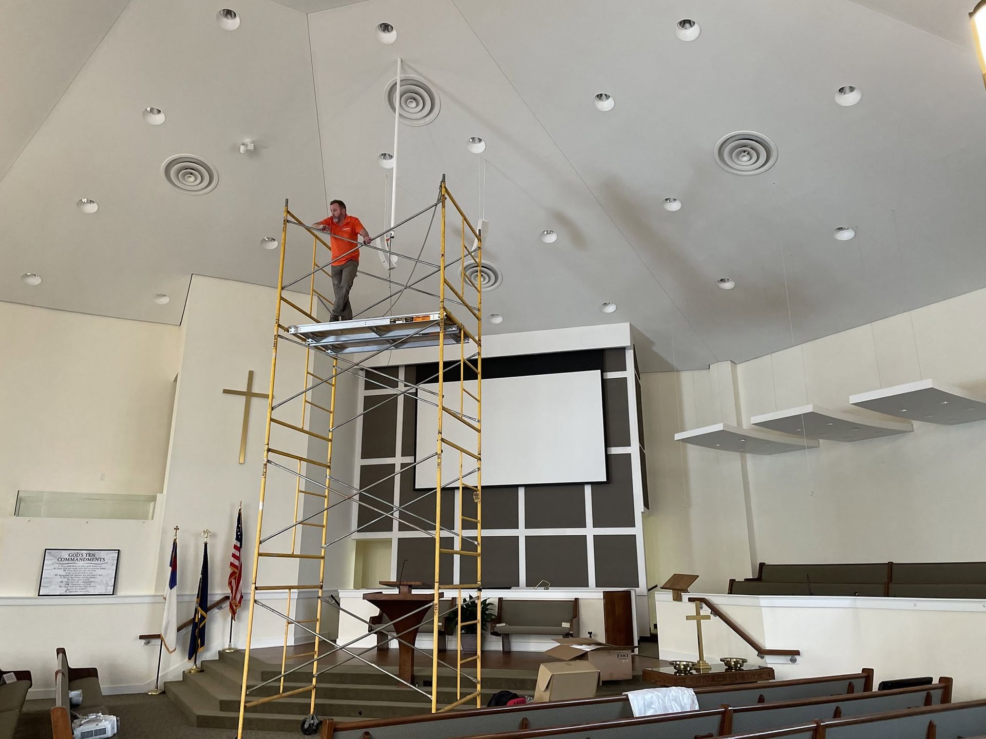 Worker on scaffolding installing equipment in a large white auditorium with rows of seats.