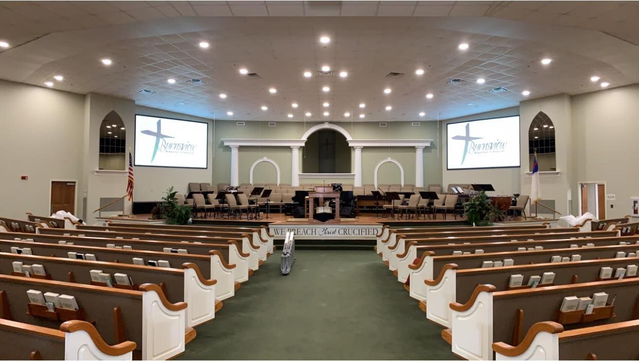 Wide church sanctuary with rows of pews facing a stage, green carpet, and two projection screens displaying a service slide