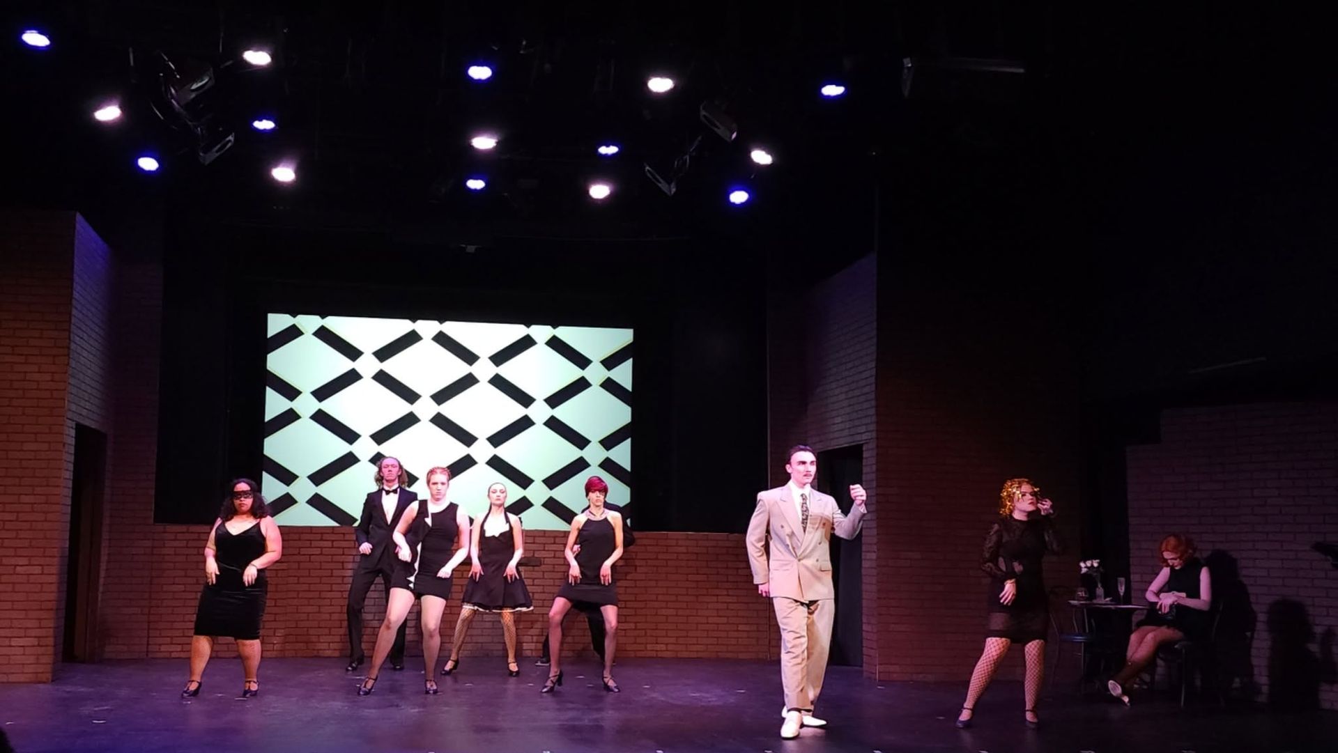 Performers on a stage with a geometric backdrop and purple lighting, arranged in a dance or theater scene