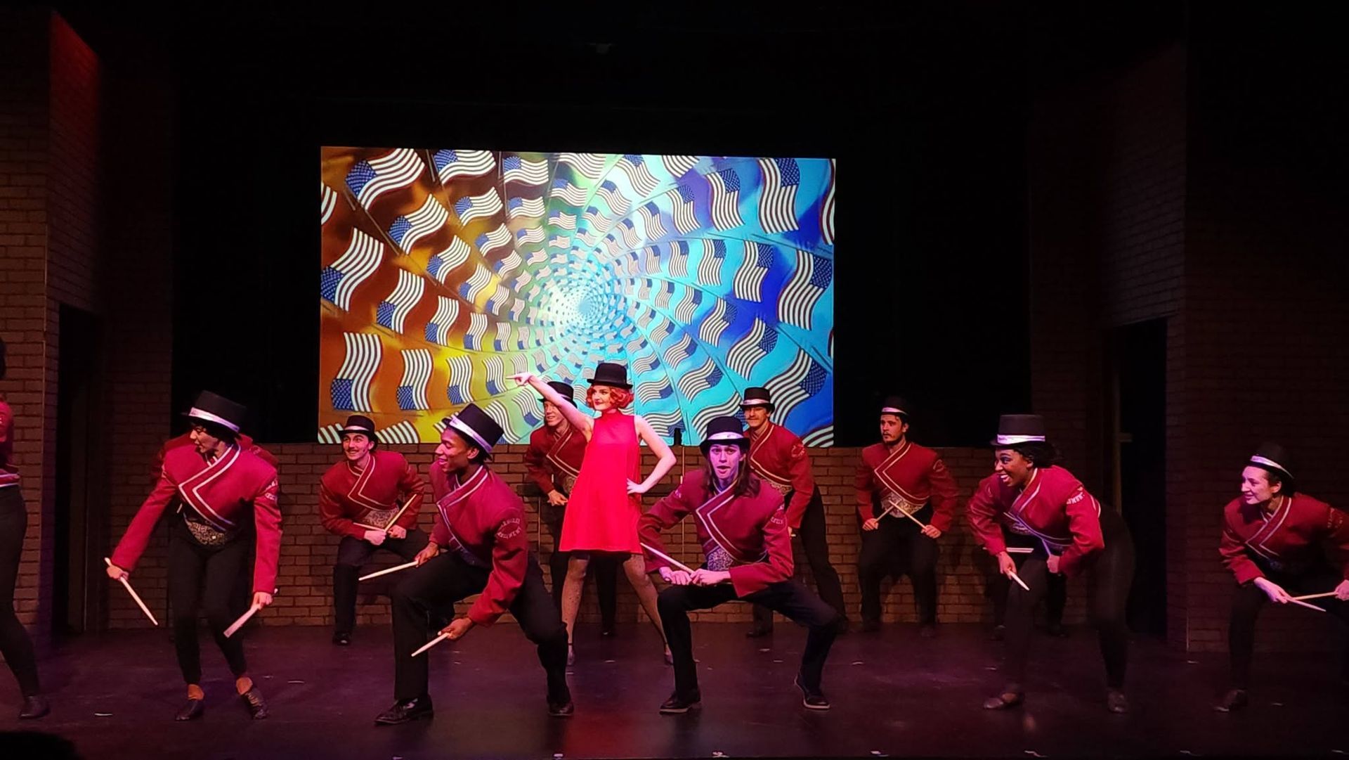 Dance troupe in red and black costumes performing on stage with a colorful abstract backdrop