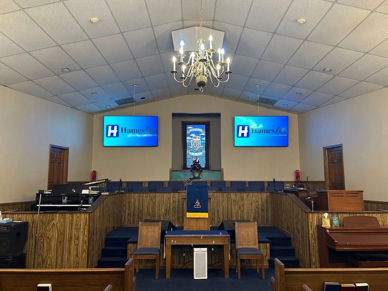 Church sanctuary stage with pulpit, chandeliers, wood paneling, and two screens displaying a logo.