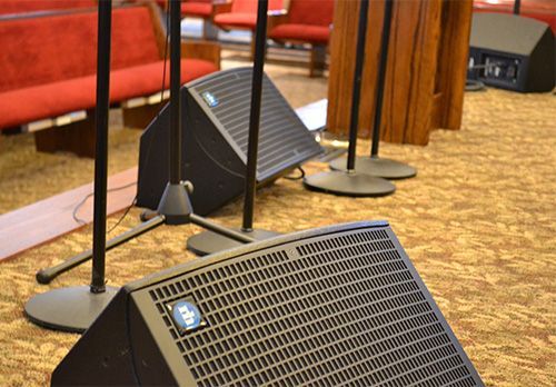Black stage speakers with cables on a carpeted floor in front of red seating and wooden columns.