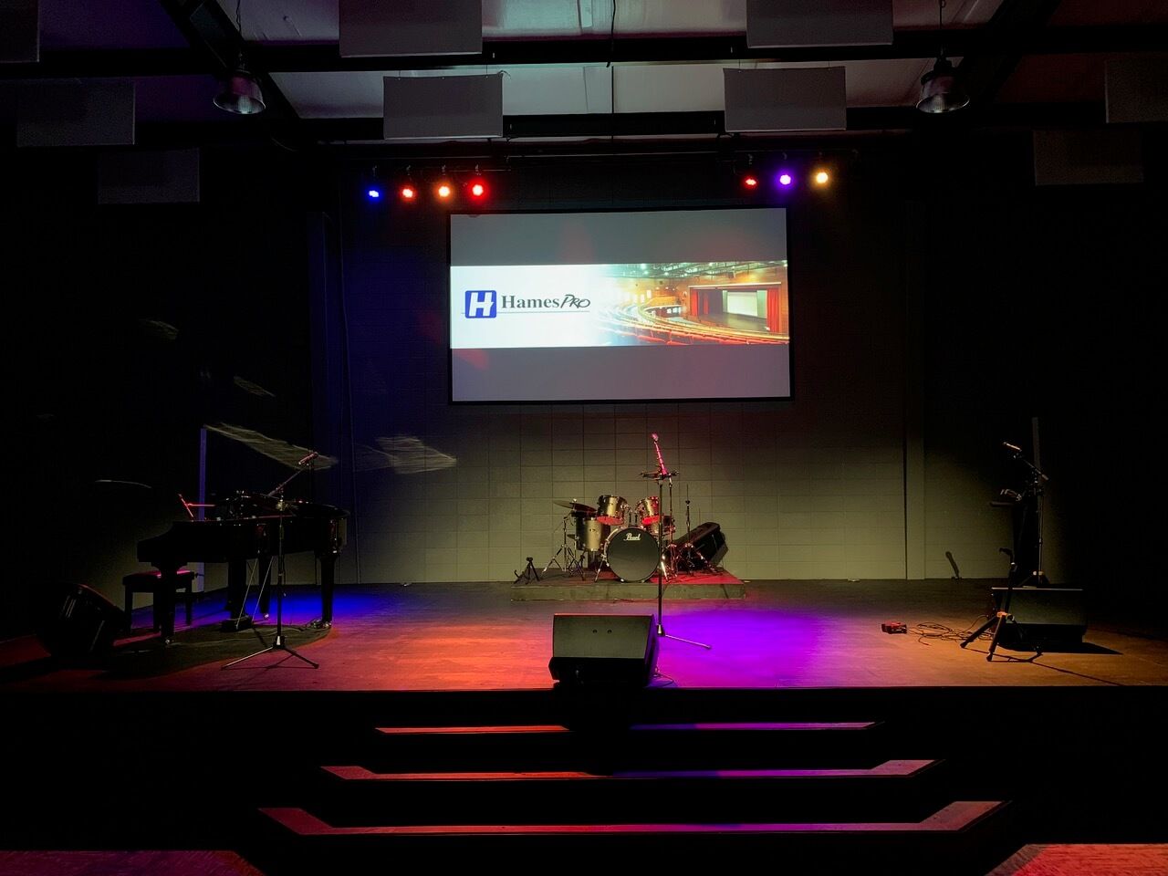Empty stage with drum kit, projector screen, and colored lights in a dark venue