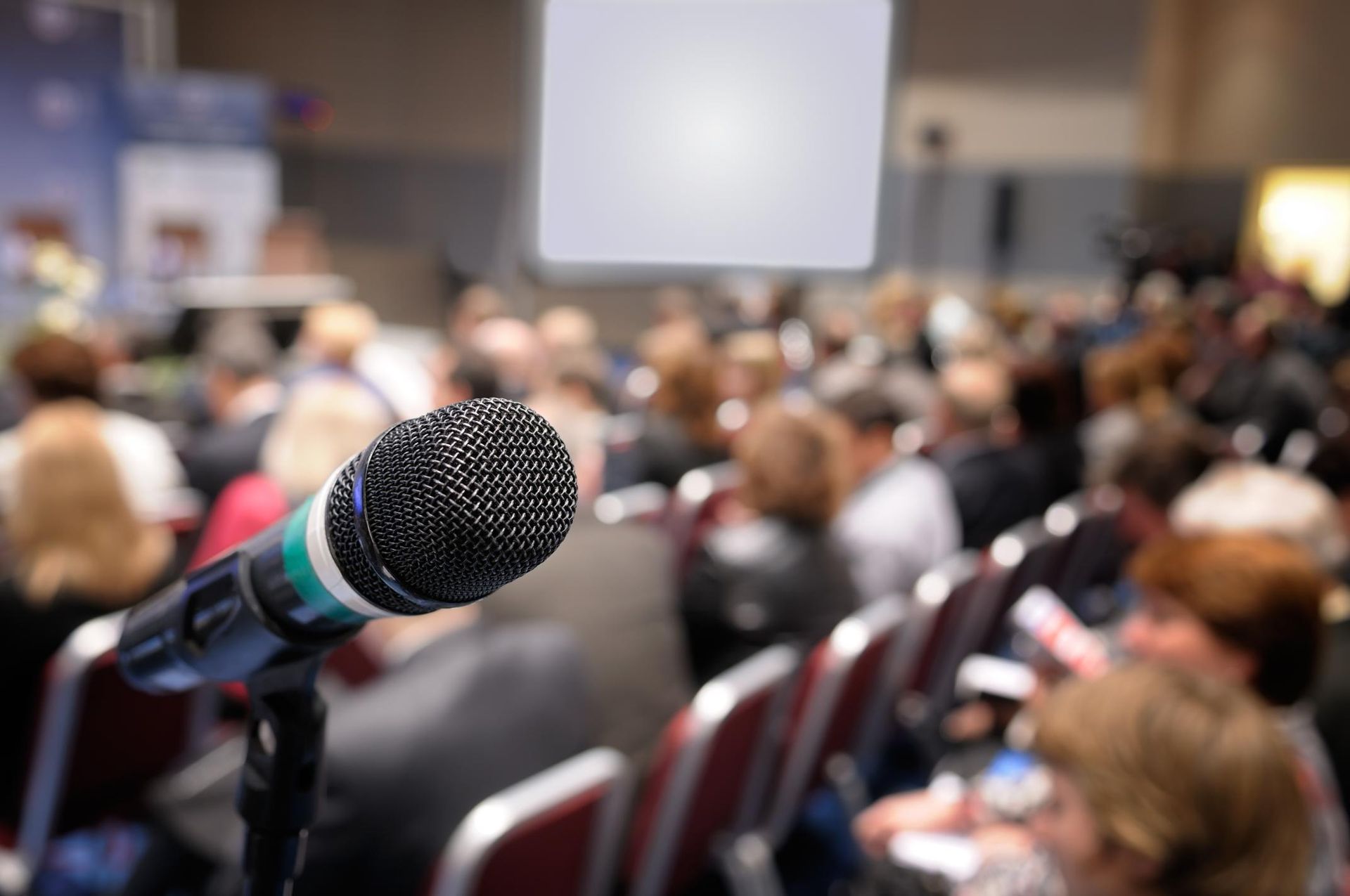 Microphone in foreground at a blurred conference audience, with a projection screen in back