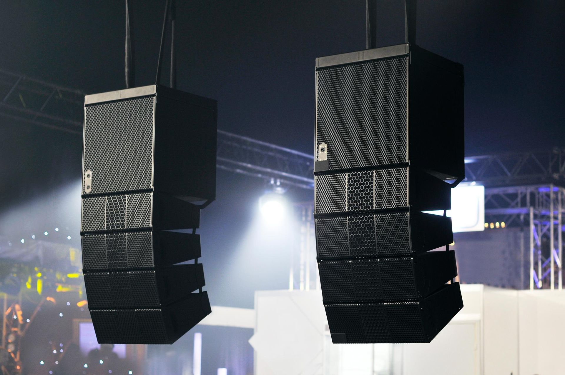 Two hanging black speaker arrays at an outdoor concert stage, lit by bright white backlights
