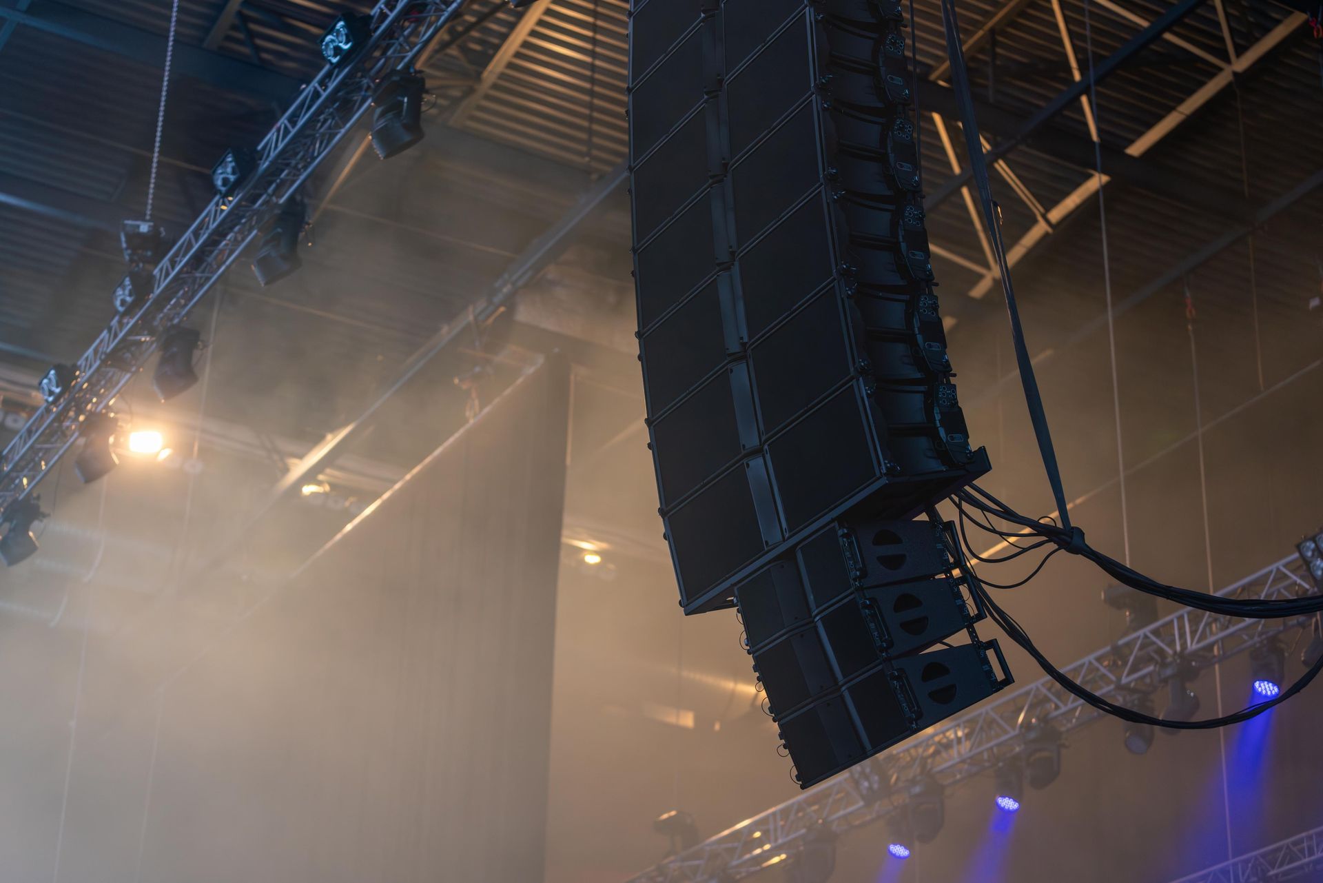 Concert stage with a large suspended speaker array, hazy smoke, and blue-purple lights overhead