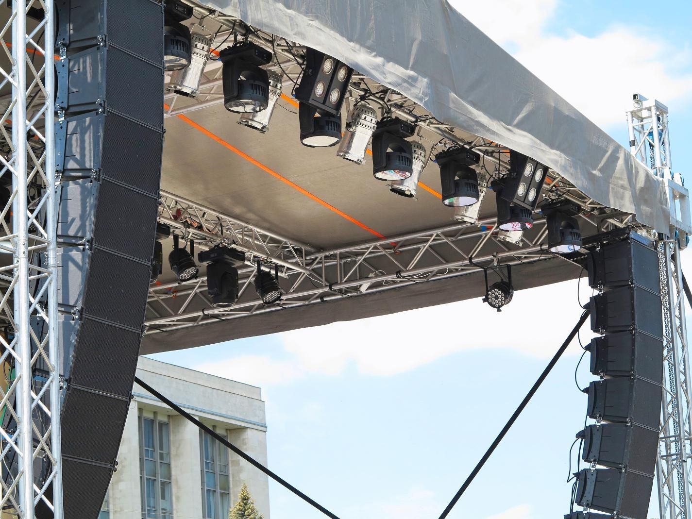 Outdoor stage truss with rows of black lights under a canopy, against a blue sky