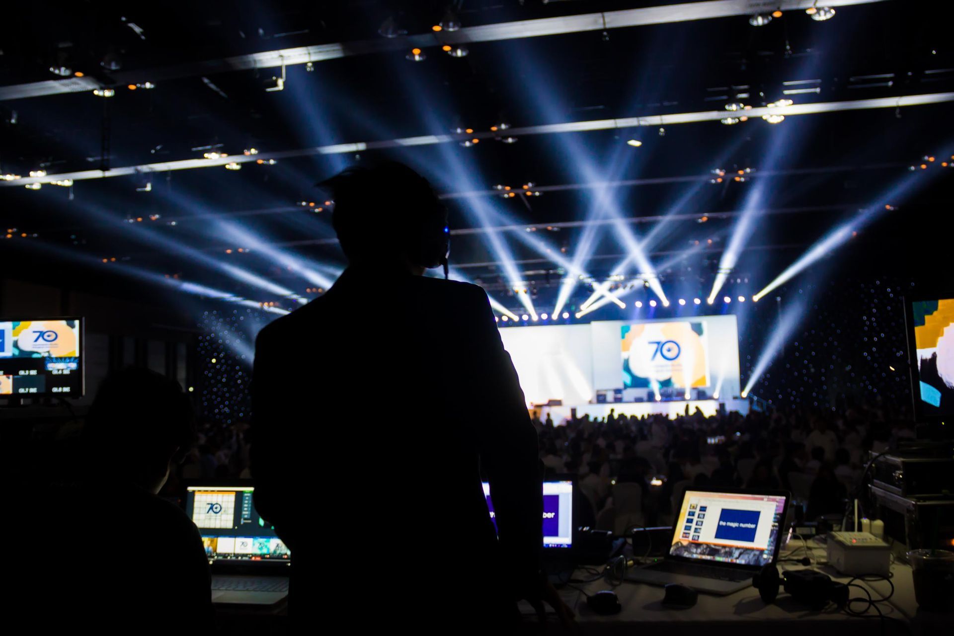 Silhouetted person in a control booth overlooking a concert stage with bright spotlights and screens