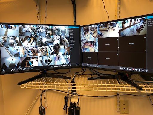 Dual computer monitors showing security camera feeds on a desk in a corner room