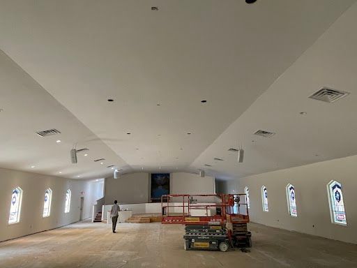 Large unfinished hall interior with white walls, arched windows, scaffolding, and construction equipment.
