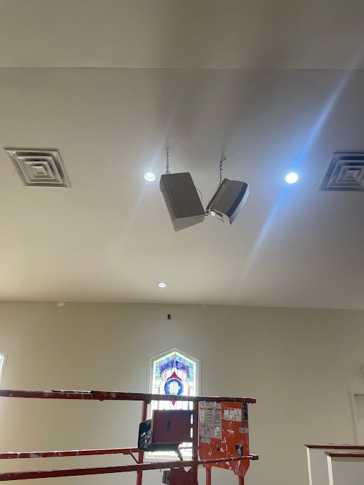 Ceiling-mounted speakers and lights above a small church or hall interior with stained-glass window below