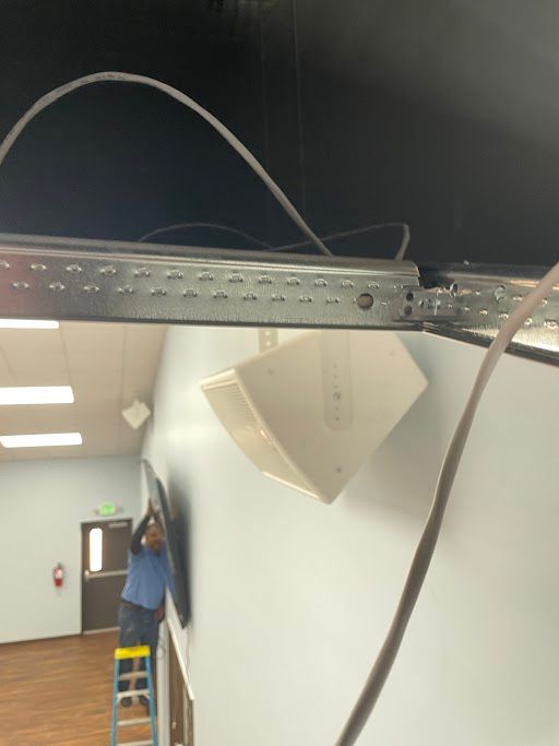 Person on a ladder working near ceiling-mounted equipment and cables in a hallway-like indoor space