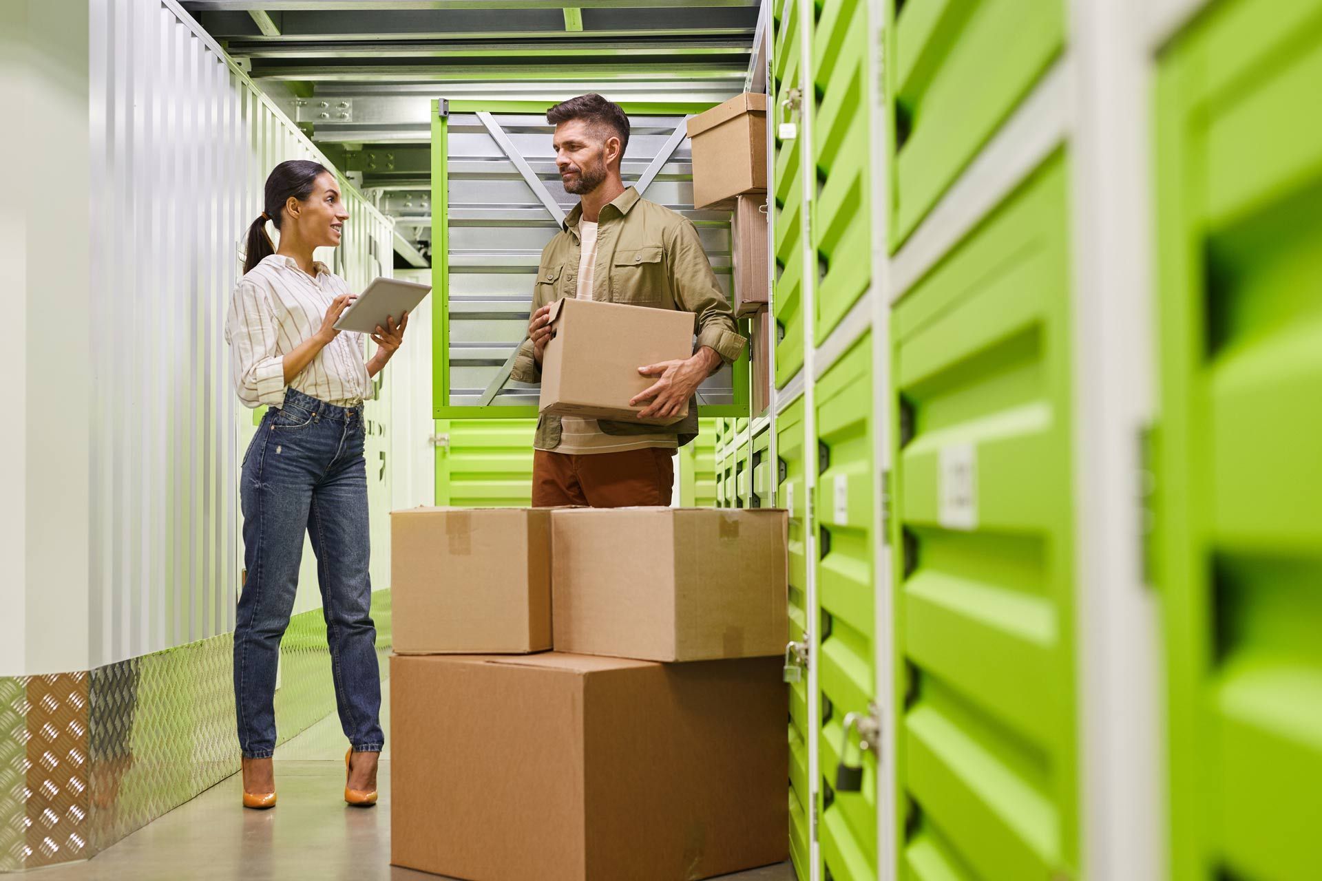 Woman with tablet, man with box in storage unit with cardboard boxes.