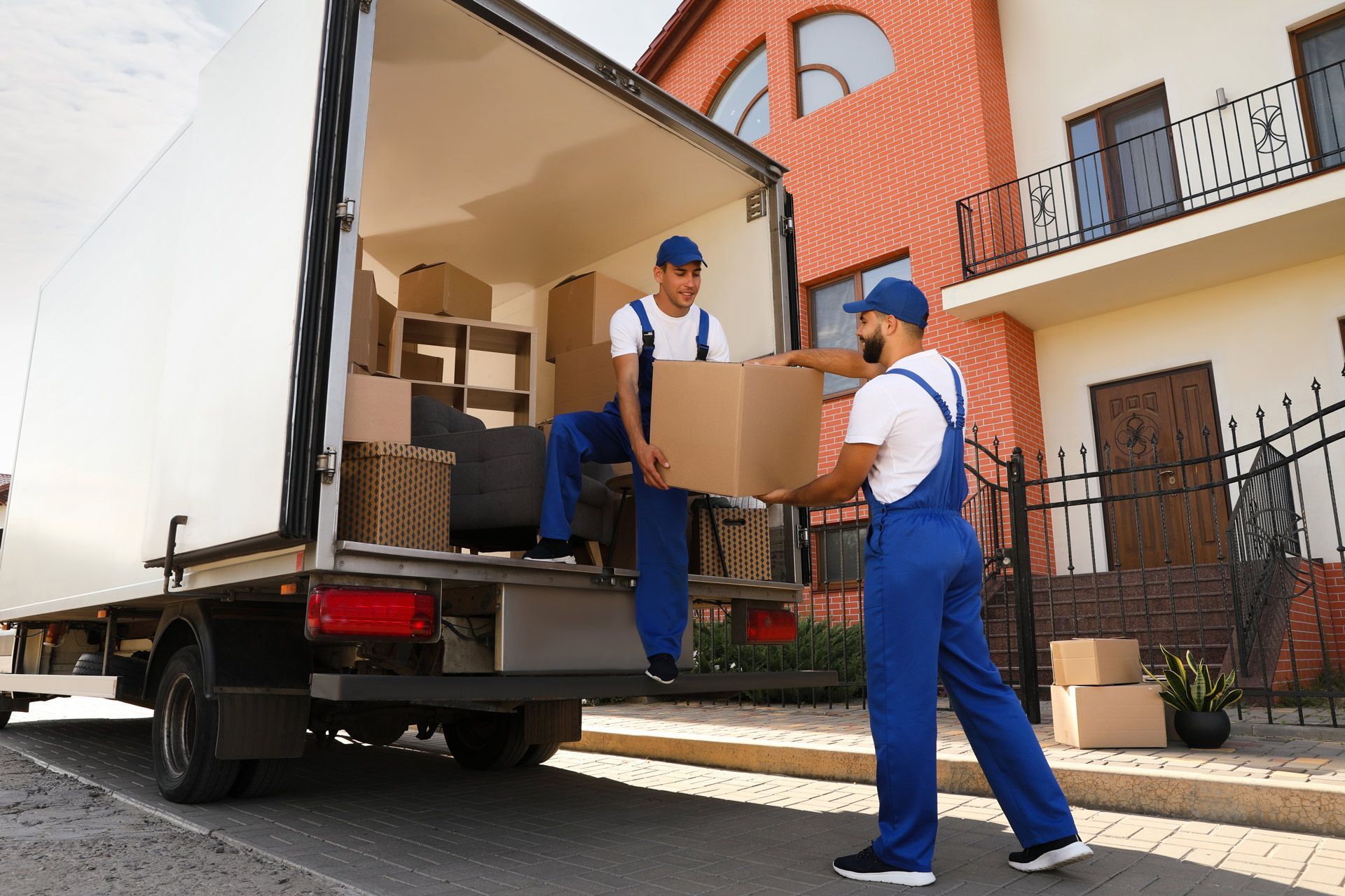 Two movers in blue uniforms unload cardboard boxes from a truck in front of a house.