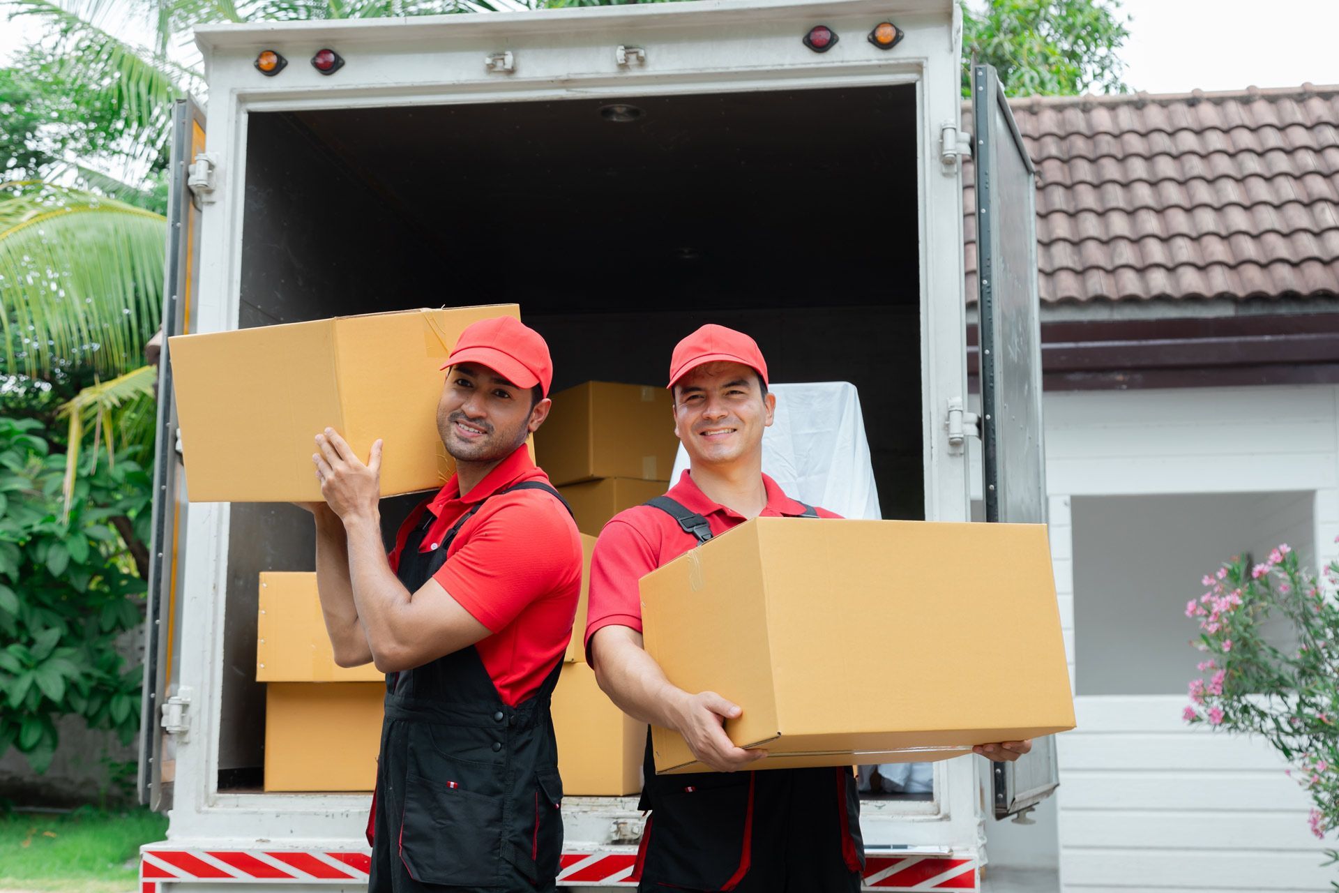 Two movers in red shirts and caps carrying cardboard boxes from a truck outside a house.