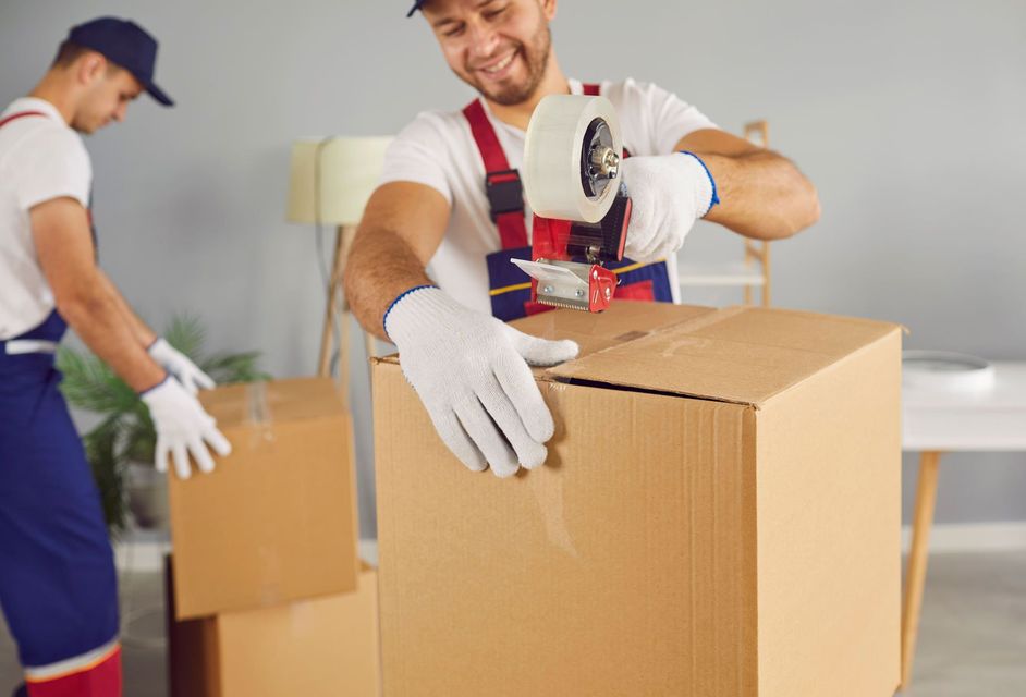 Two movers packing boxes, one taping a box, smiling. Indoors, gray walls.