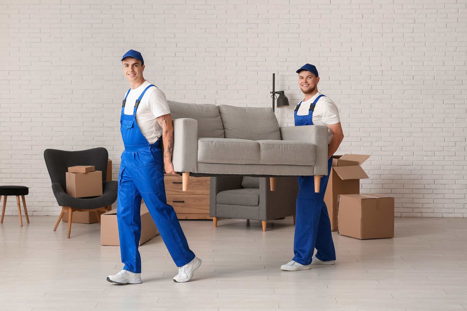 Two movers in blue overalls carrying a gray sofa in a room with boxes, smiling.