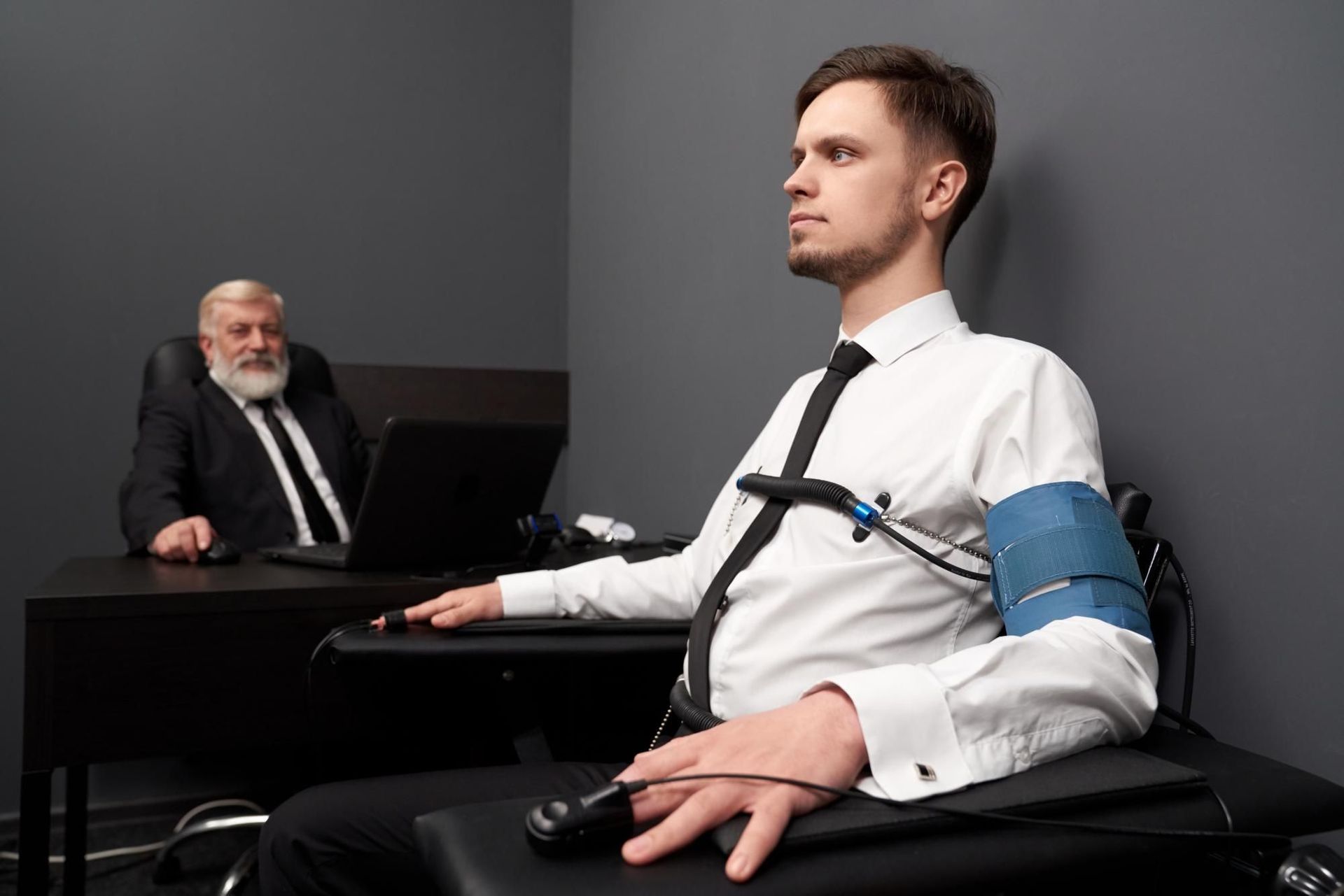 Man taking a polygraph test, seated with equipment attached. Older man with laptop in background.