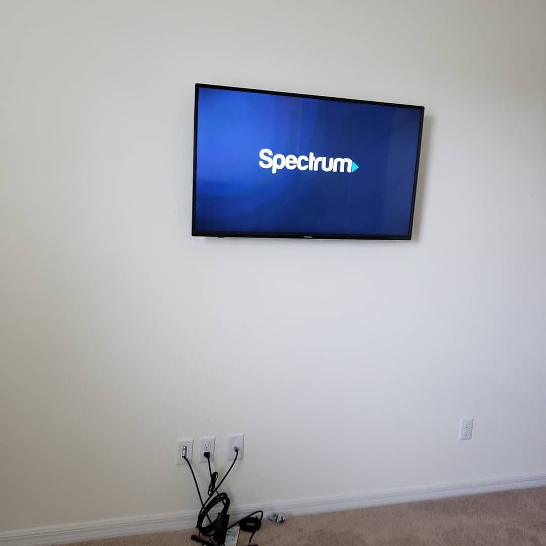 A wall-mounted TV displaying the Spectrum logo. Power cords and outlets are visible below. Beige walls.