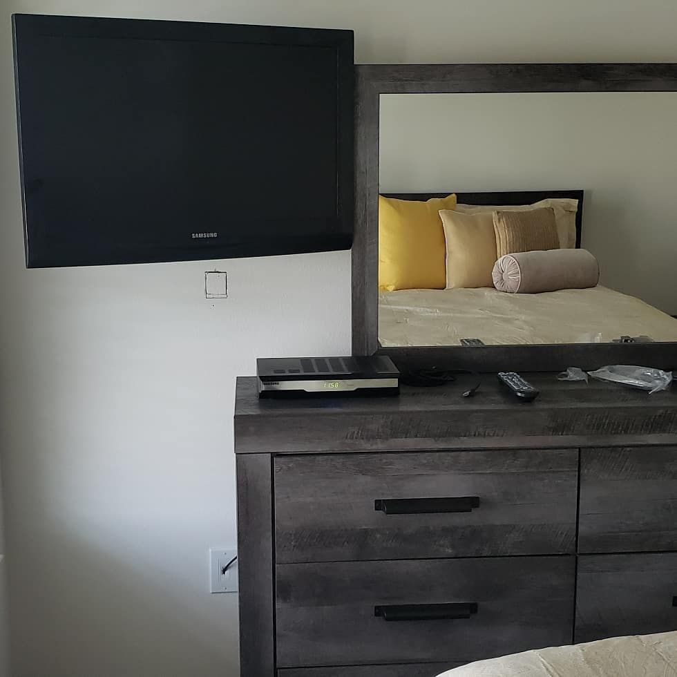 A bedroom dresser with mirror. A TV is mounted on the wall above the dresser. The bed is reflected in the mirror.