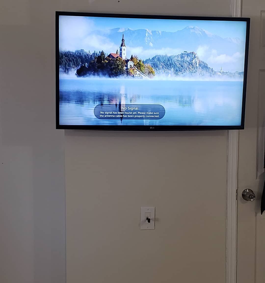 TV mounted on a light-colored wall, displaying a lake scene. Below, a small electrical outlet is visible.