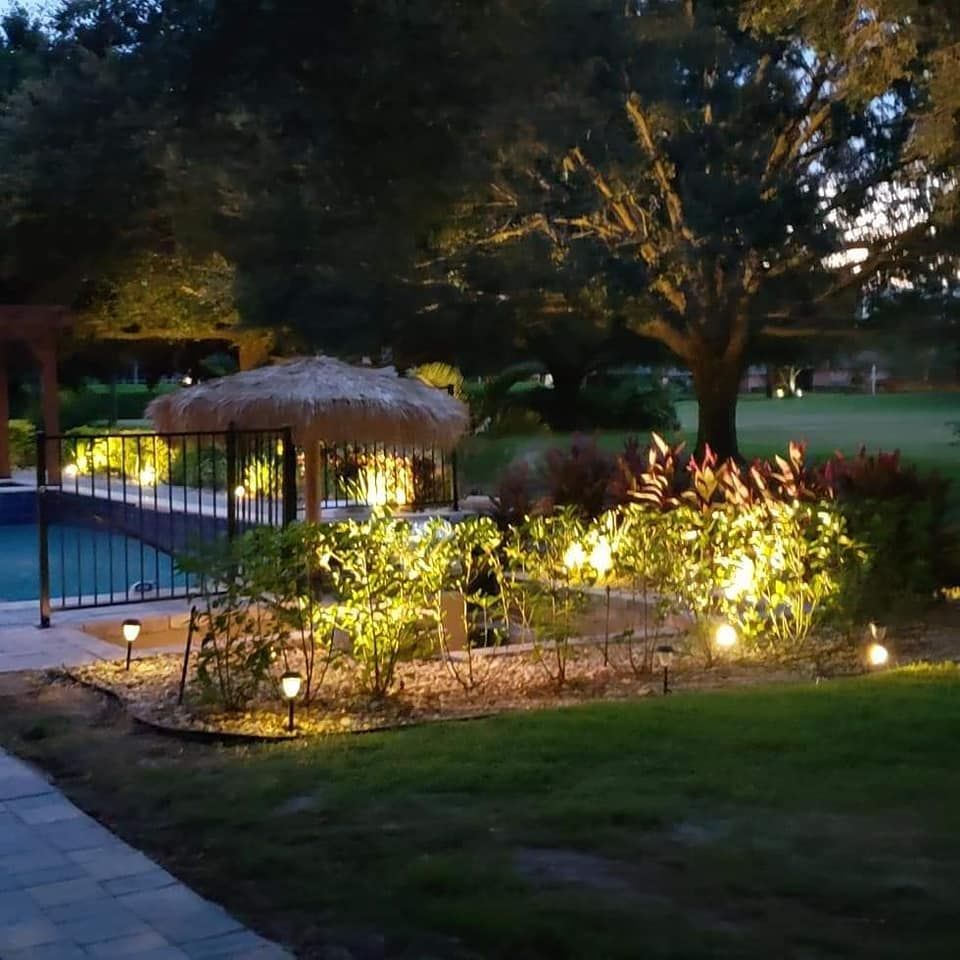 Lit landscaping around a pool and thatched roof cabana at dusk.
